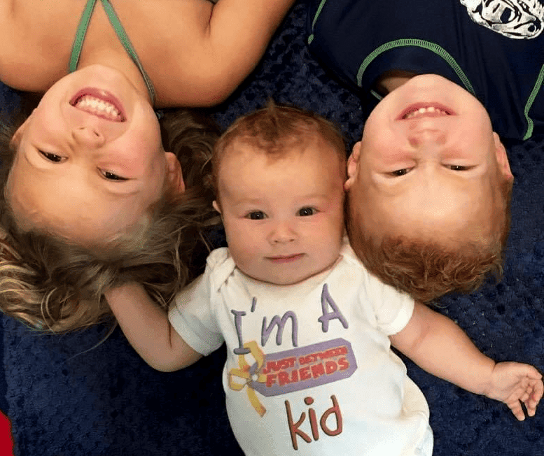 Three children lying on the floor looking up.  The baby has a onesie on that says, "I'm a Just Between Friends Kid"