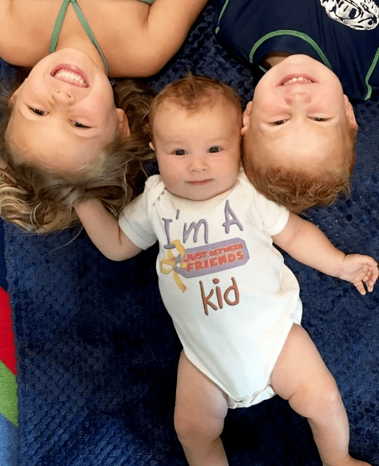 Three children lying on the floor looking up.  The baby has a onesie on that says, "I'm a Just Between Friends Kid"